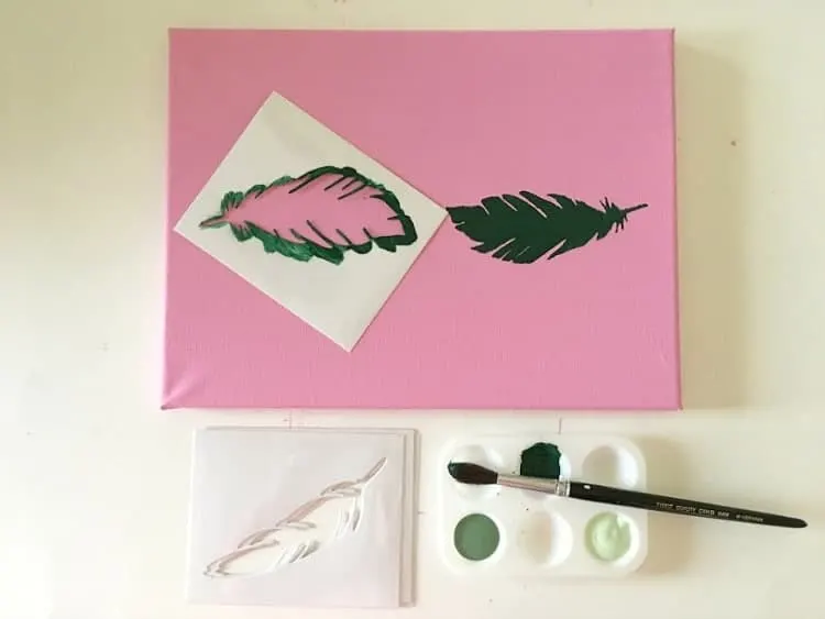 Using a stencil to paint a green feather on a pink background.