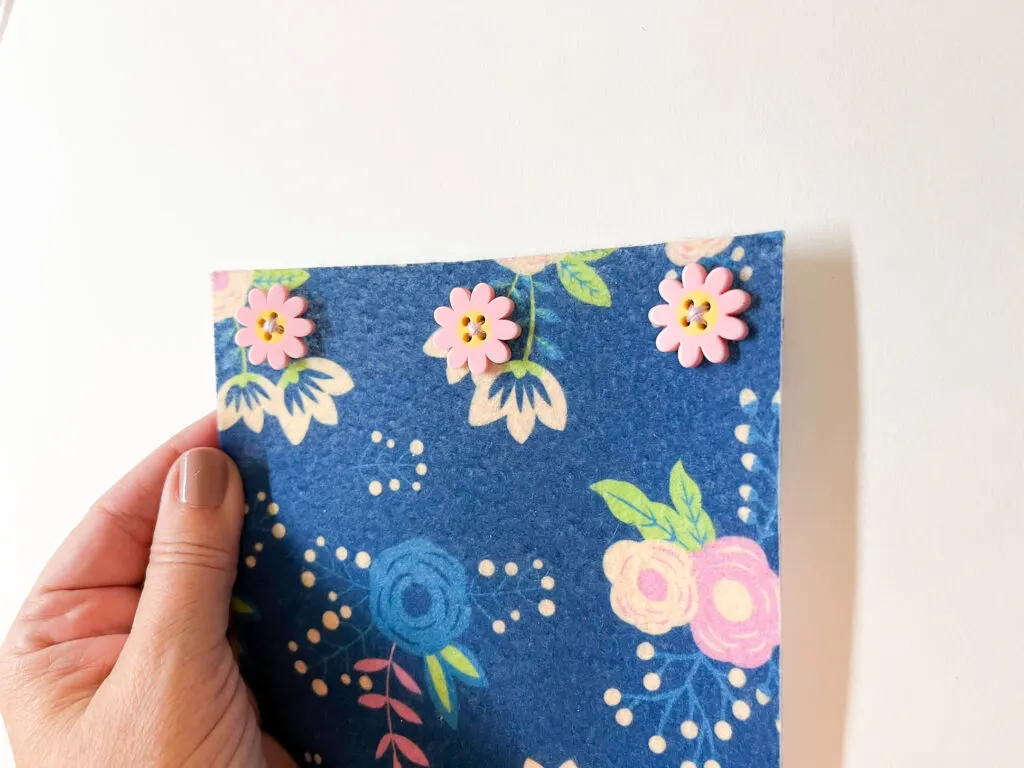 Three pink daisy buttons sewn along the edge of a piece of blue floral fabric to make a french press cozy.