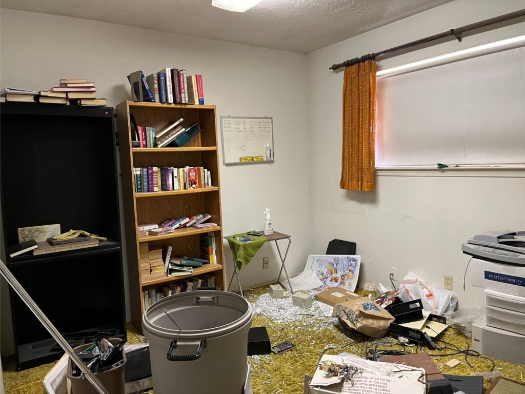 A cluttered messy room with green shag carpet and orange curtains.