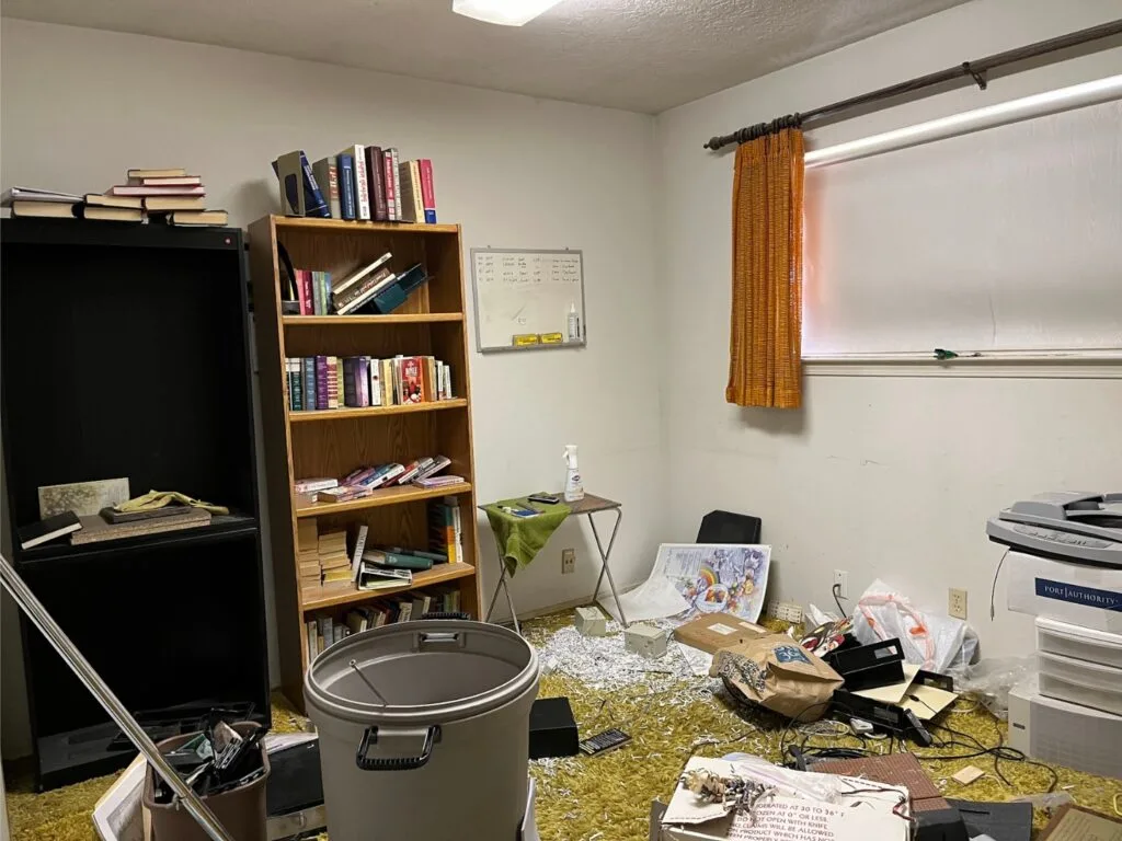 A cluttered messy room with green shag carpet and orange curtains.