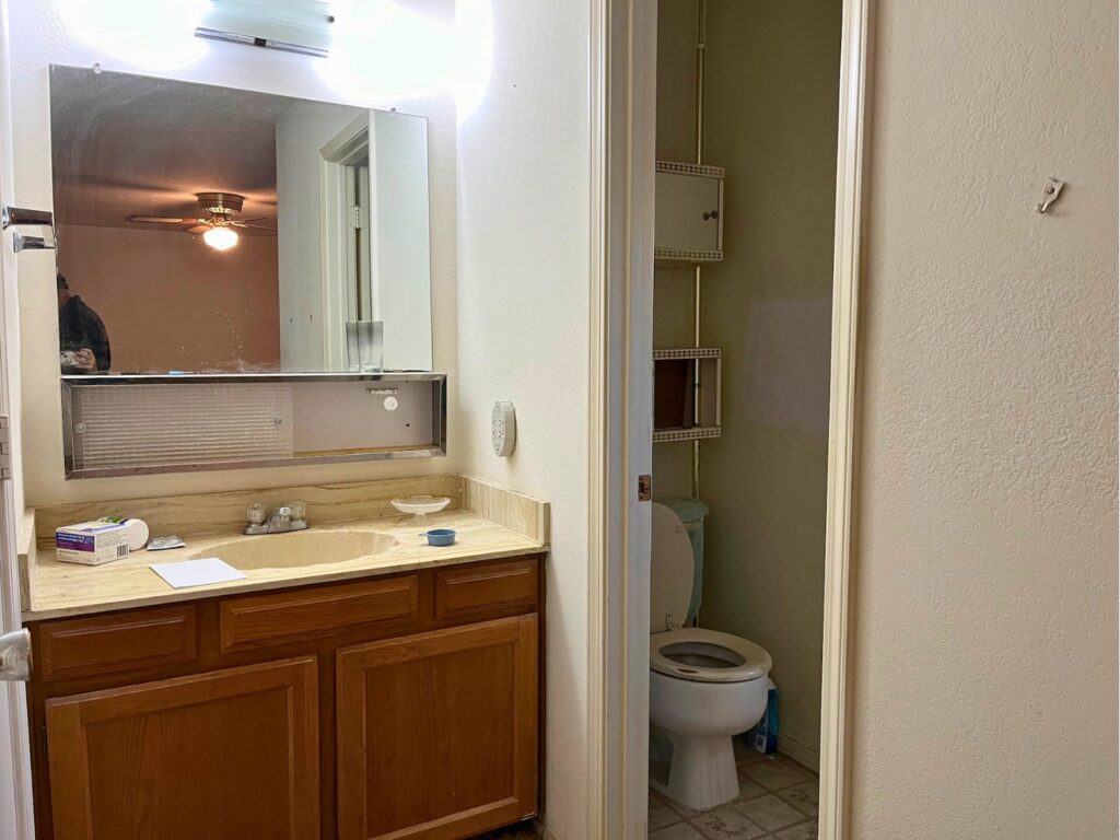 A dated bathroom sink area, with a glimpse of the toilet and shower room through the doorway.