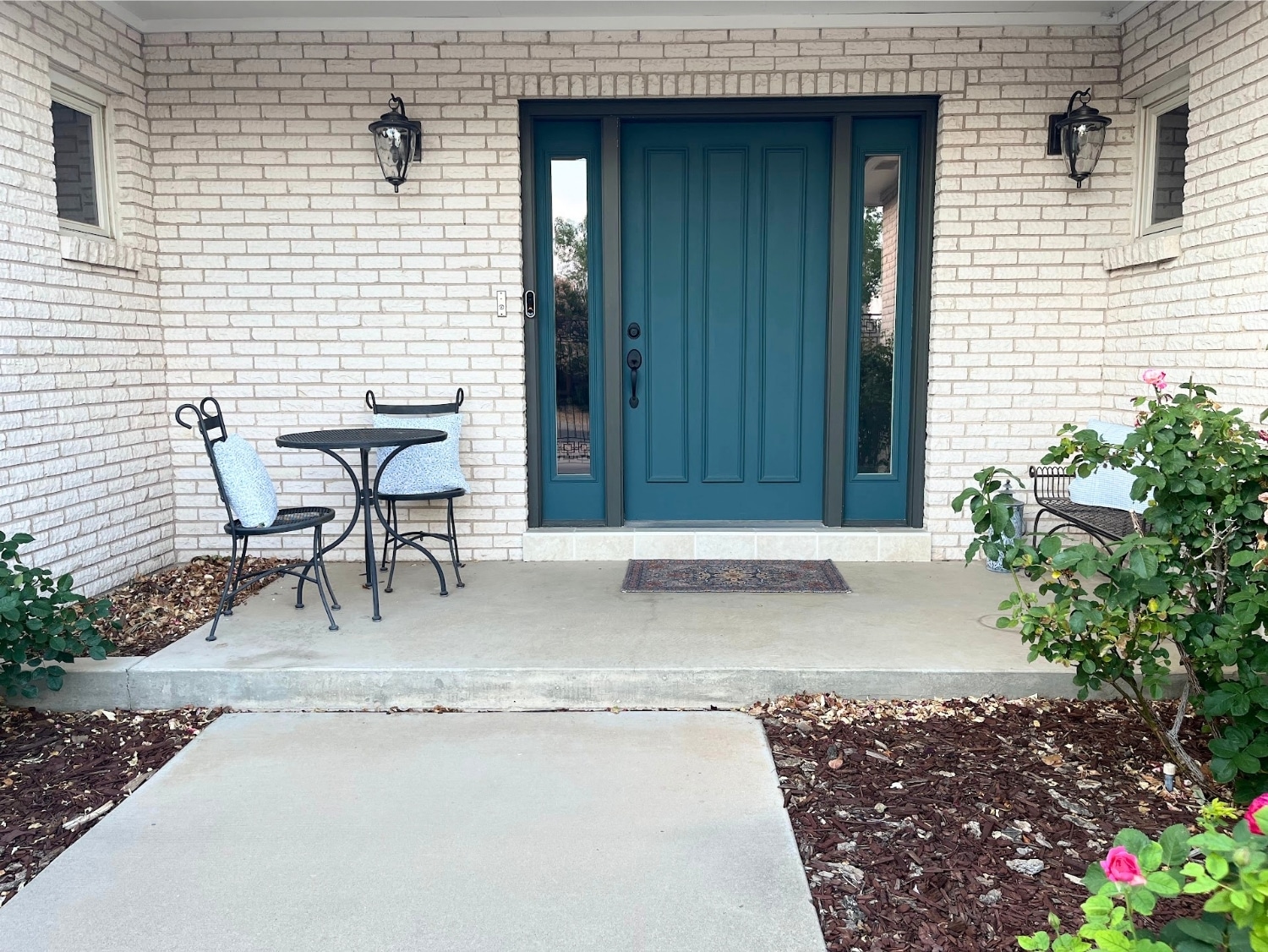Front entryway of a white brick house with dark teal front door and black wrought iron bistro set.